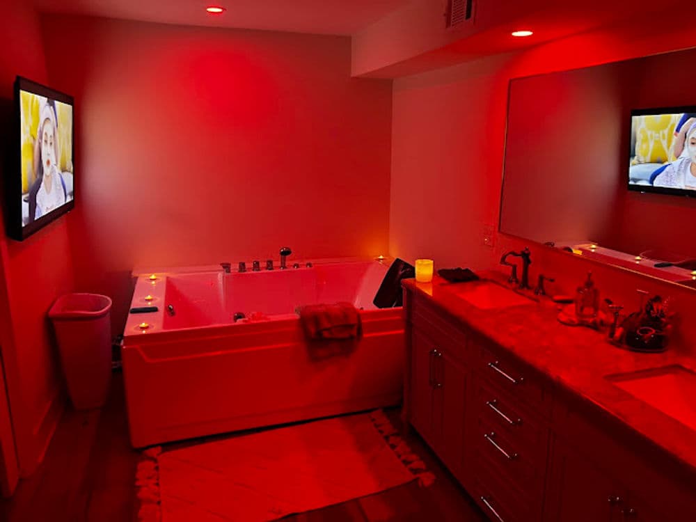 Luxurious bathroom with red lighting, a jetted tub, wall-mounted TV, and elegant decor.