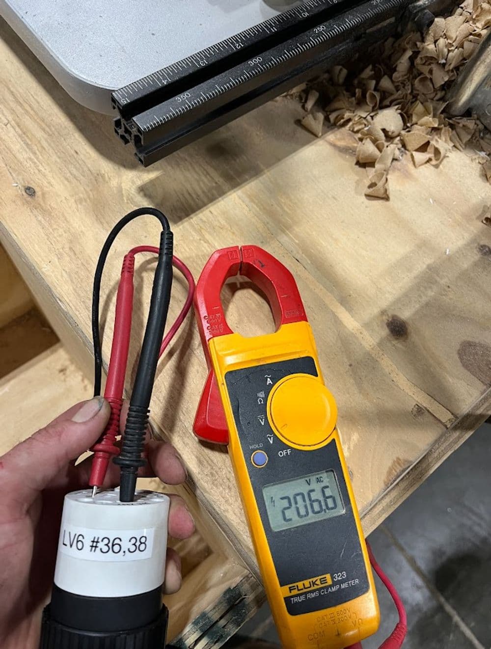 User holding a Fluke clamp meter displaying voltage on a workbench with wood shavings.