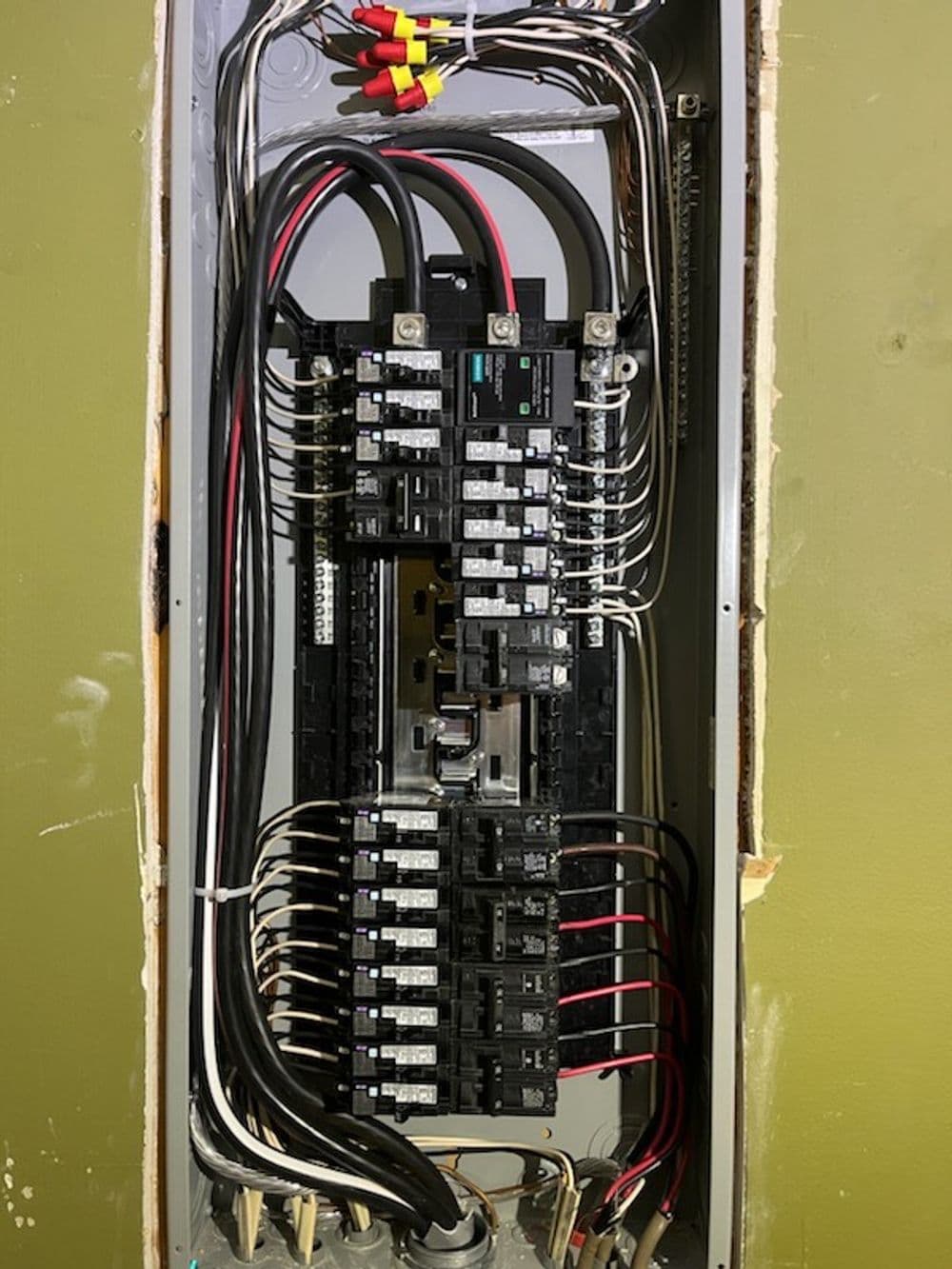Electrical panel with circuit breakers and wiring mounted on a green wall.