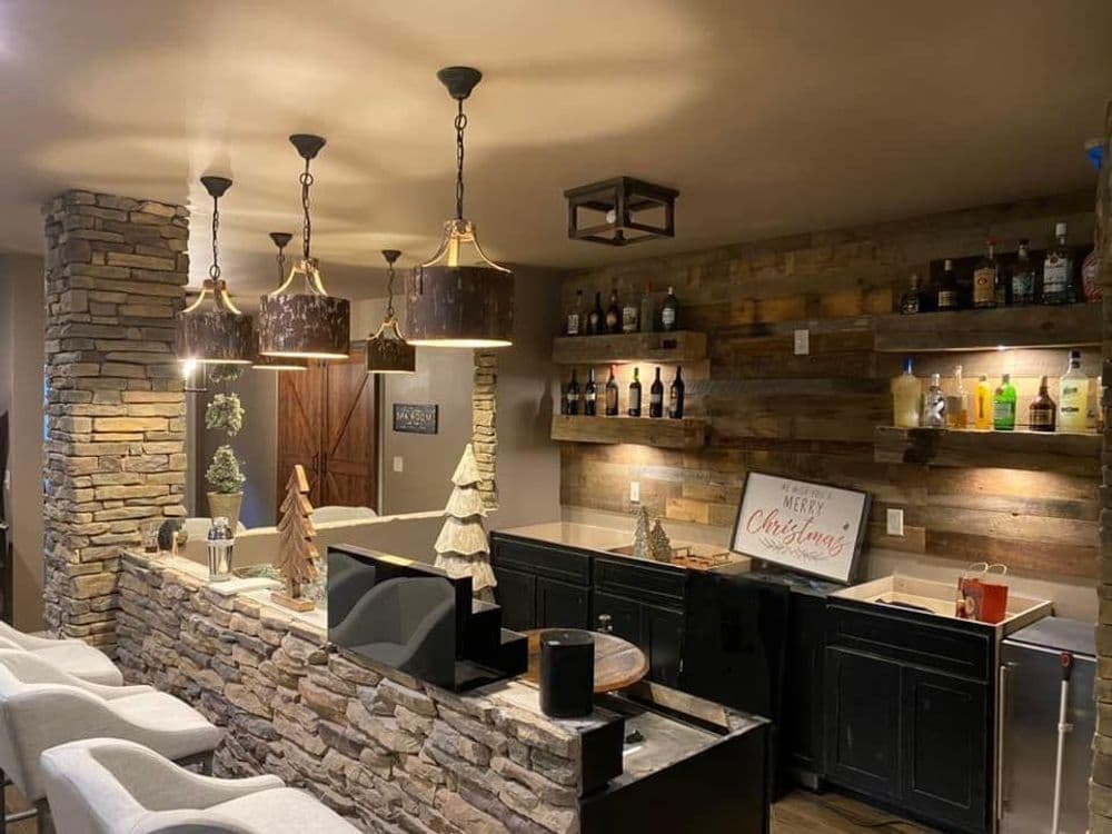 Cozy home bar with stone accents, wooden shelves, festive decor, and stylish pendant lights.