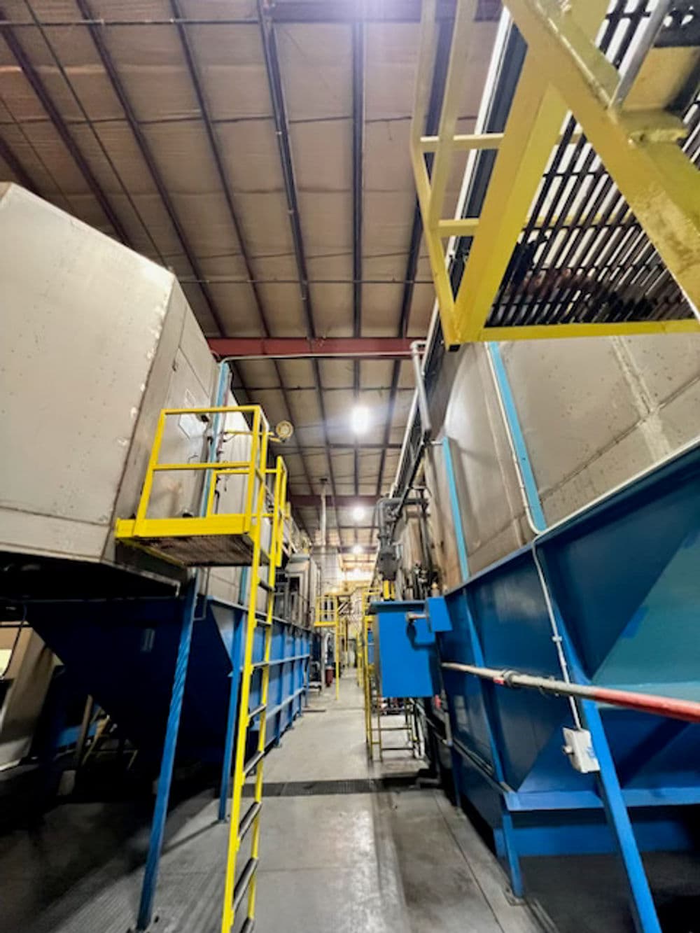 Industrial facility interior with large machinery, metal stairs, and overhead lighting.