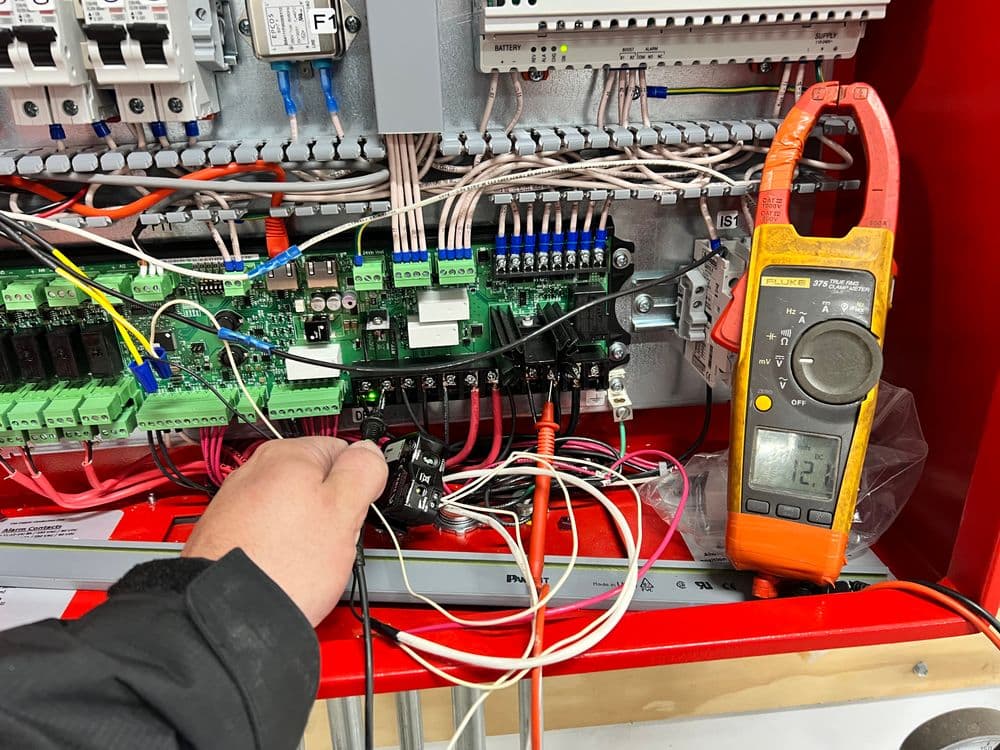 Electrician troubleshooting circuit board with multimeter and wiring in control panel.