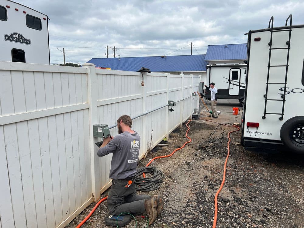 Electricians installing outdoor outlets along a white fence at a commercial site.