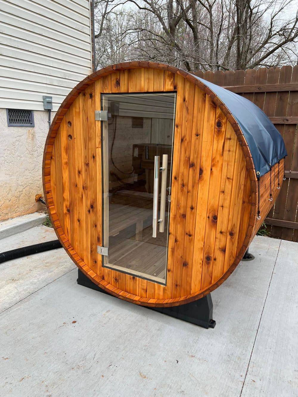 Modern wooden barrel sauna with glass door, located in a backyard setting.
