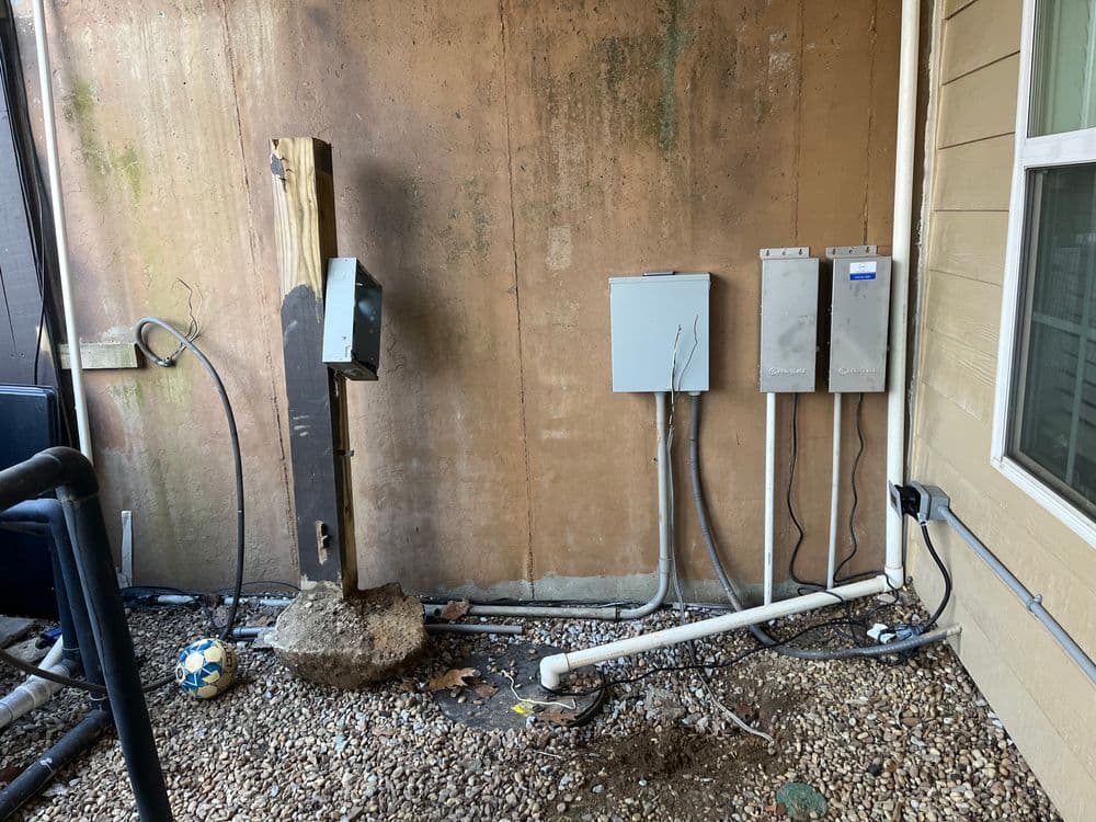 Utility equipment on an exterior wall with conduit, cables, and a wooden post in a residential setting.