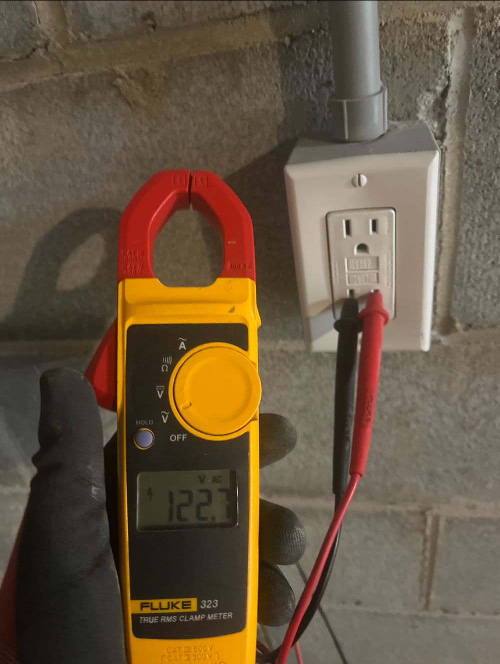Fluke 323 clamp meter measuring 122.7 volts at a wall outlet with test leads connected.