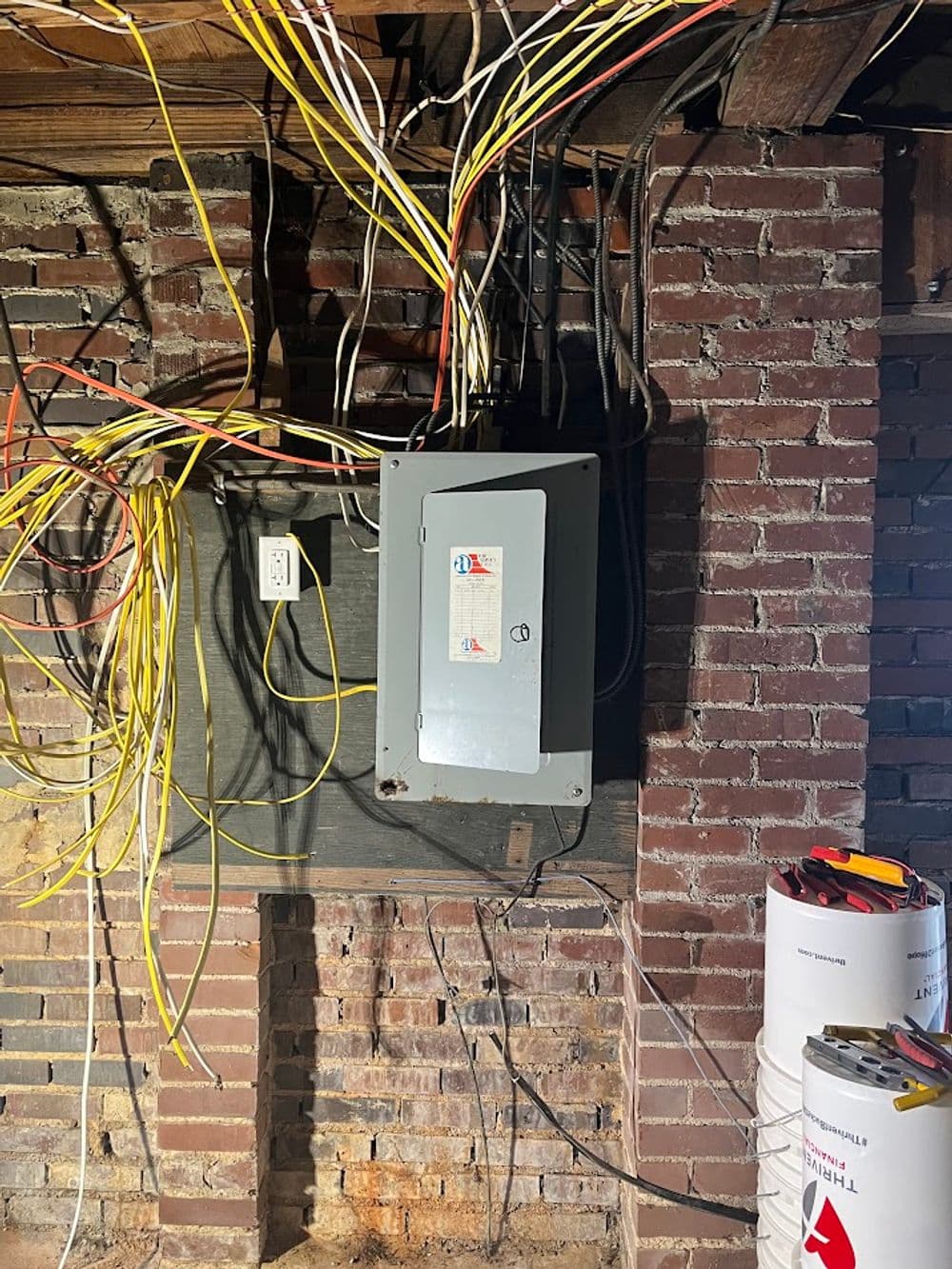 Electrical panel surrounded by tangled wires in a basement with exposed brick wall.