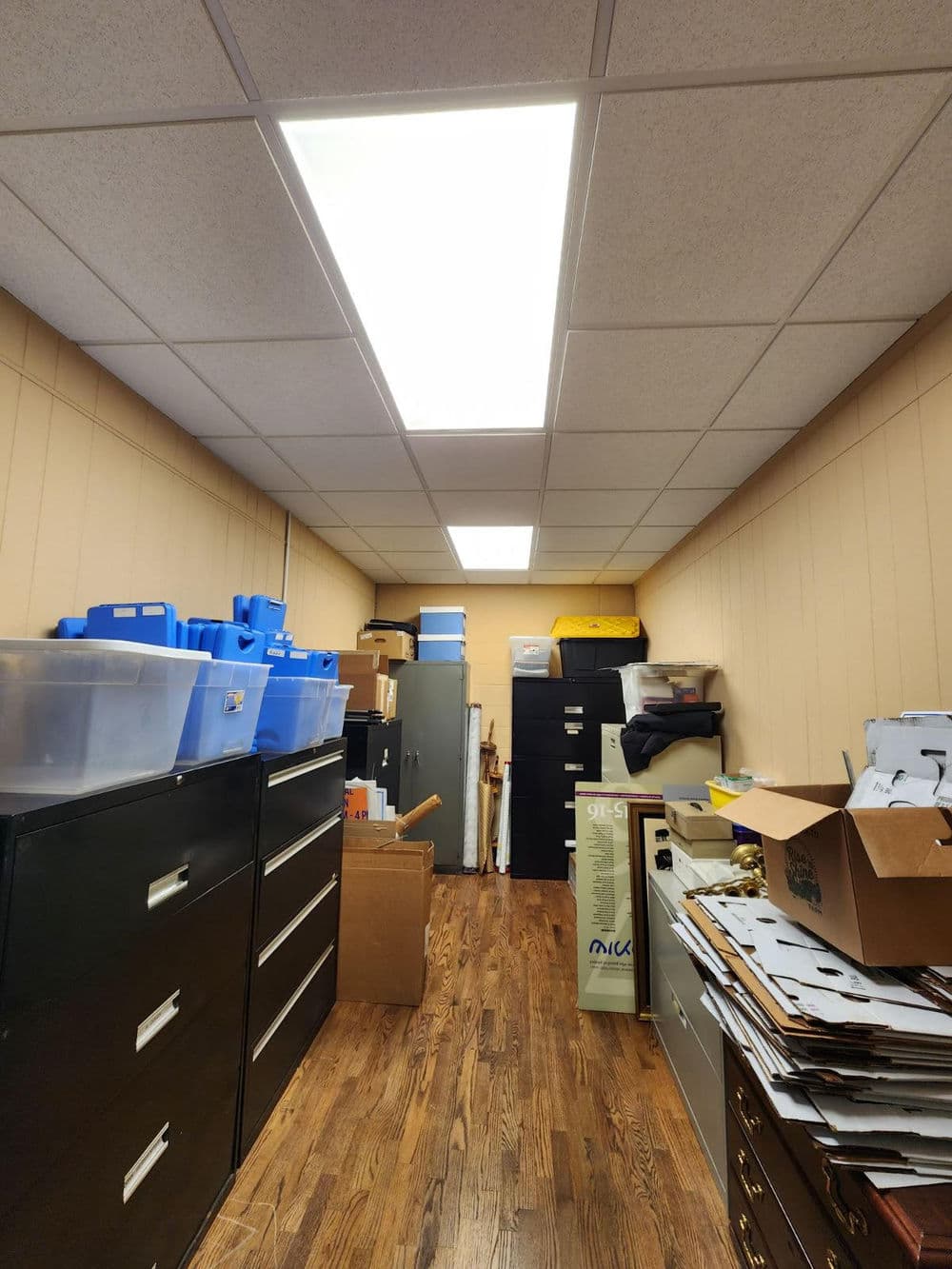 Storage area with filing cabinets, blue bins, and boxes in a cluttered office space.