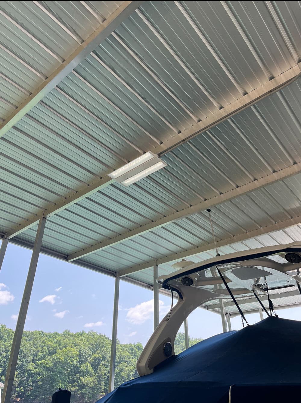 Boat covered in blue tarp under metal canopy with clear sky and trees in the background.