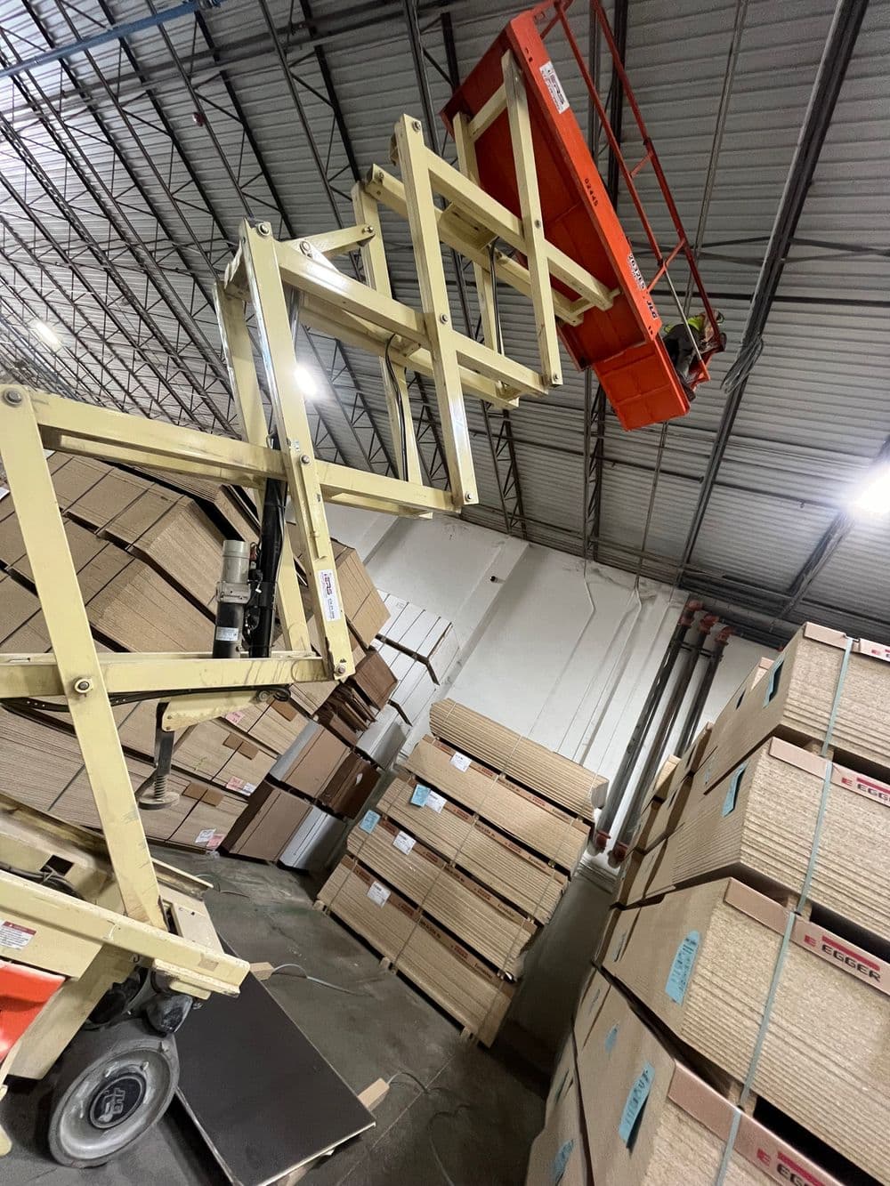 Aerial work platform lifting in a warehouse stacked with cardboard boxes.